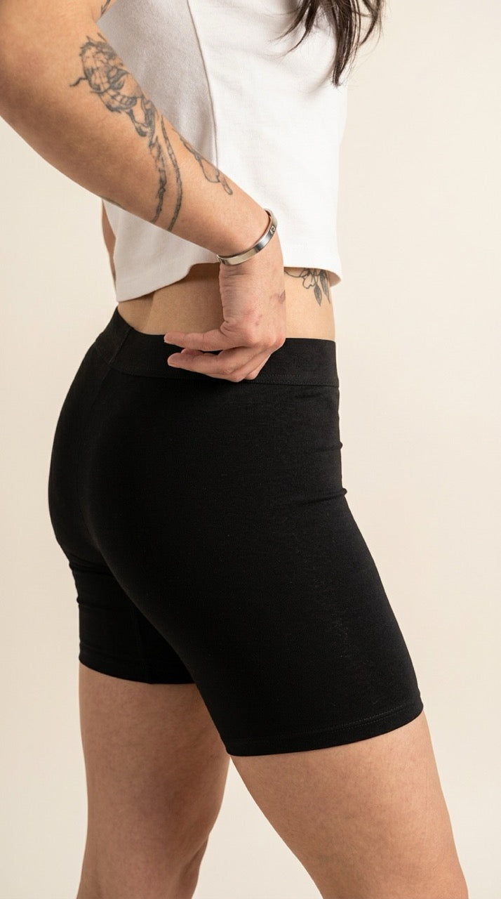 A close-up side view of a person wearing black fitted biker shorts with a white cropped top, standing against a plain light beige background.