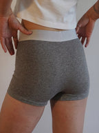 A person wearing heather gray boxer briefs with a contrasting light gray waistband, shown from the back with hands pulling the waistband slightly outward.