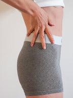 A close-up of a person wearing a heather gray high-waisted brief or shapewear shorts with a contrasting white waistband, with their hand resting on the hip area.