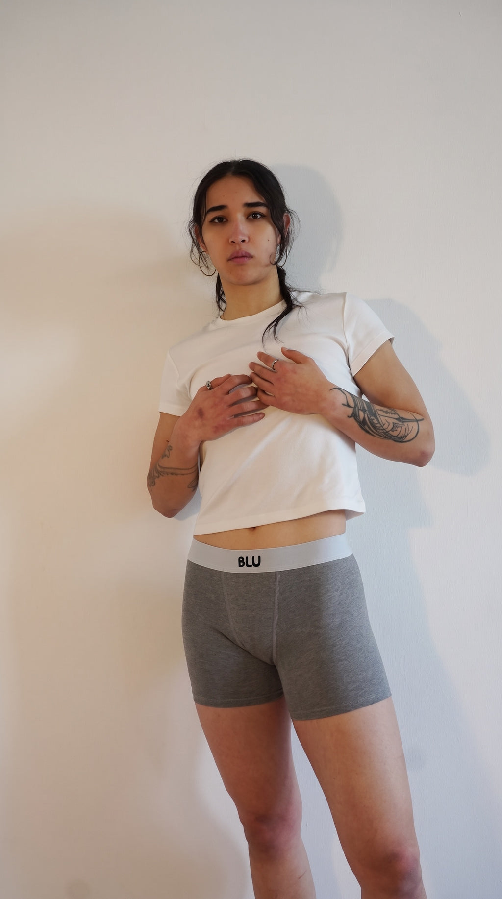 A woman modeling a pair of gray heathered boxer briefs with a white waistband that has the brand name 'BLU' printed on it, paired with a white cropped t-shirt, standing against a plain off-white wall.