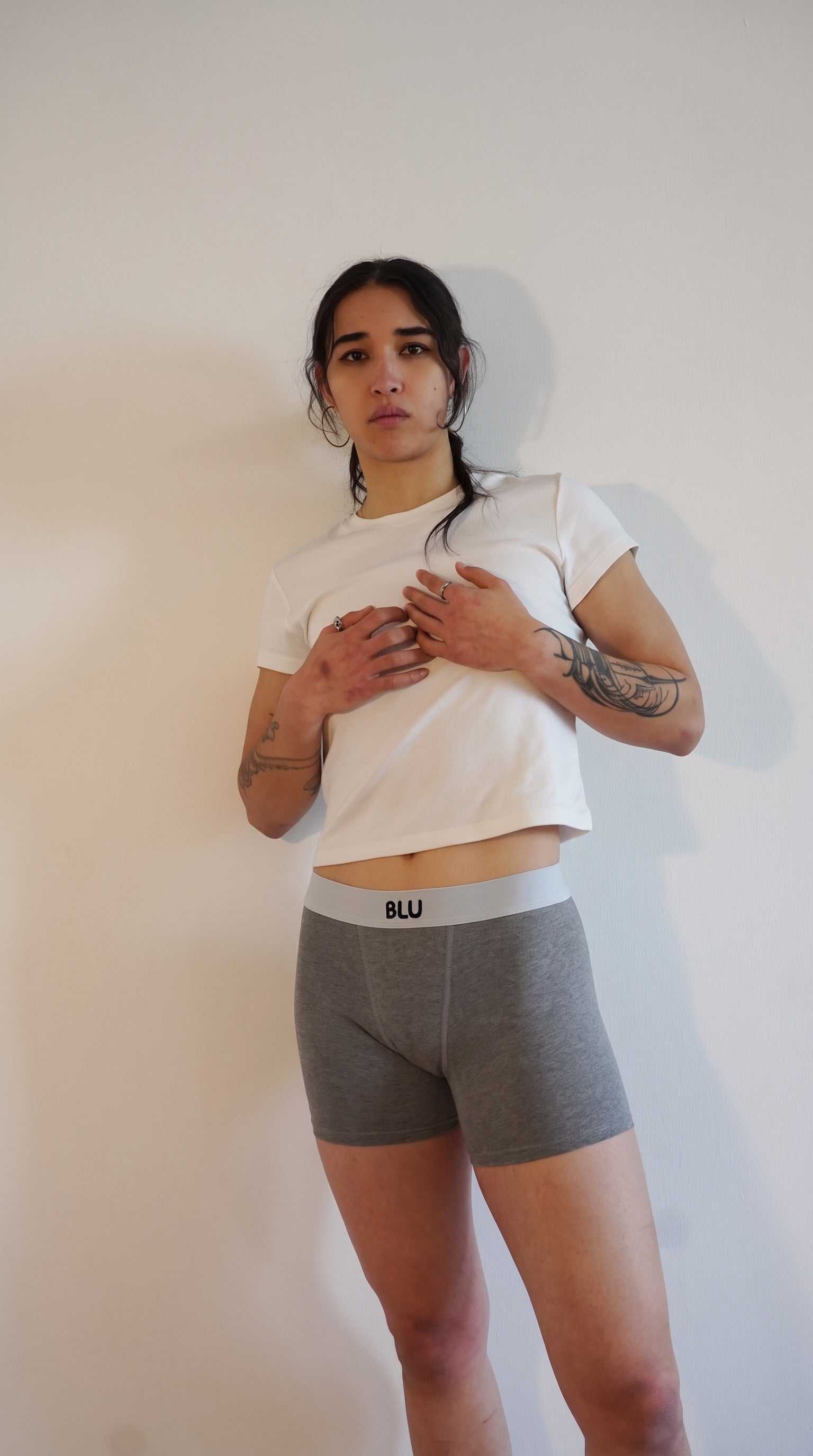 A woman modeling a pair of gray heathered boxer briefs with a white waistband that has the brand name 'BLU' printed on it, paired with a white cropped t-shirt, standing against a plain off-white wall.